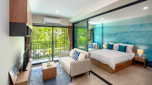 A modern hotel room with a large bed, a sofa, a TV, and a balcony with a view of greenery.