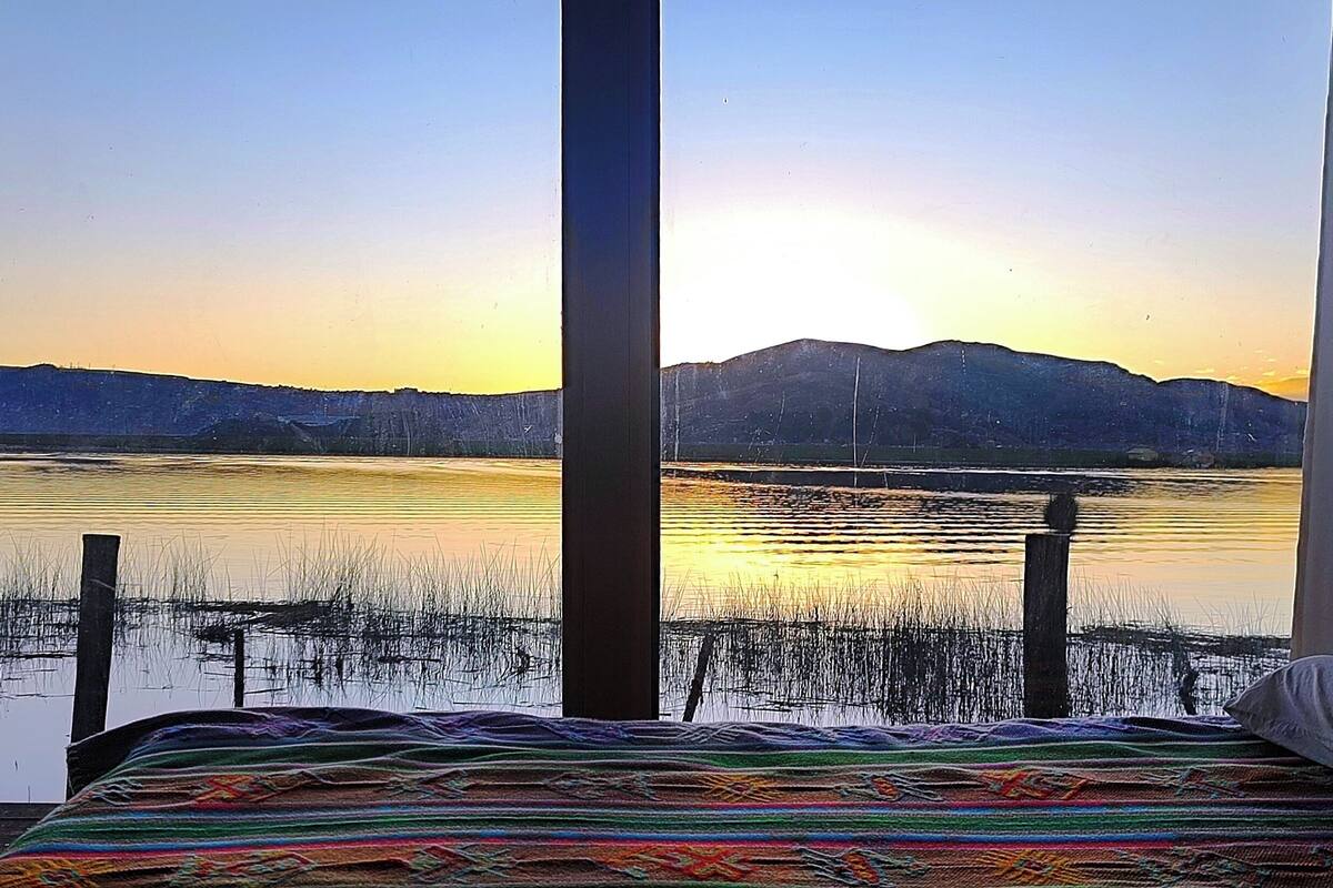 A room with a large window overlooking a lake at sunset, a bed with a colorful blanket, and a view of distant mountains.