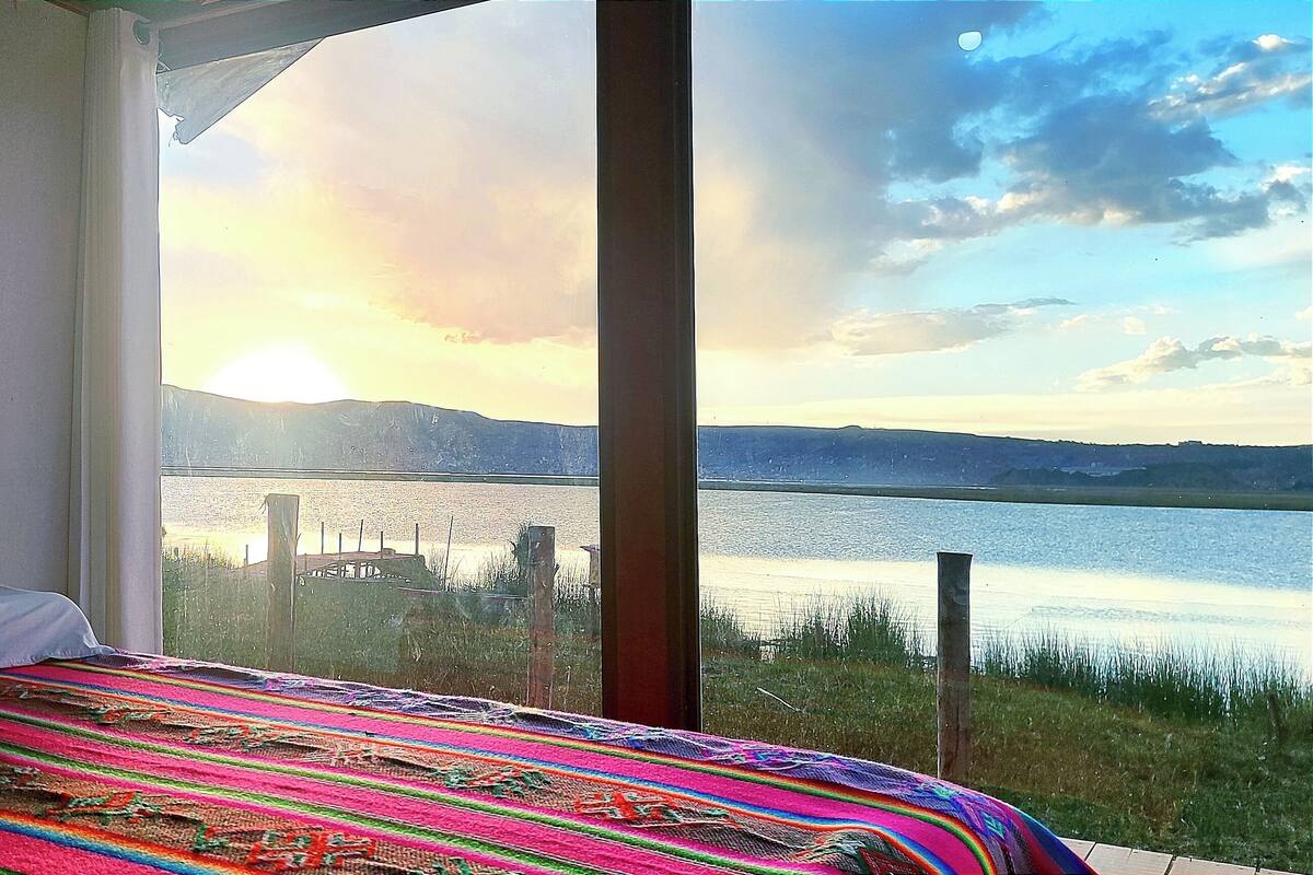A bedroom with a large window overlooking a lake, a colorful bedspread, and a lotus flower artwork on the wall.
