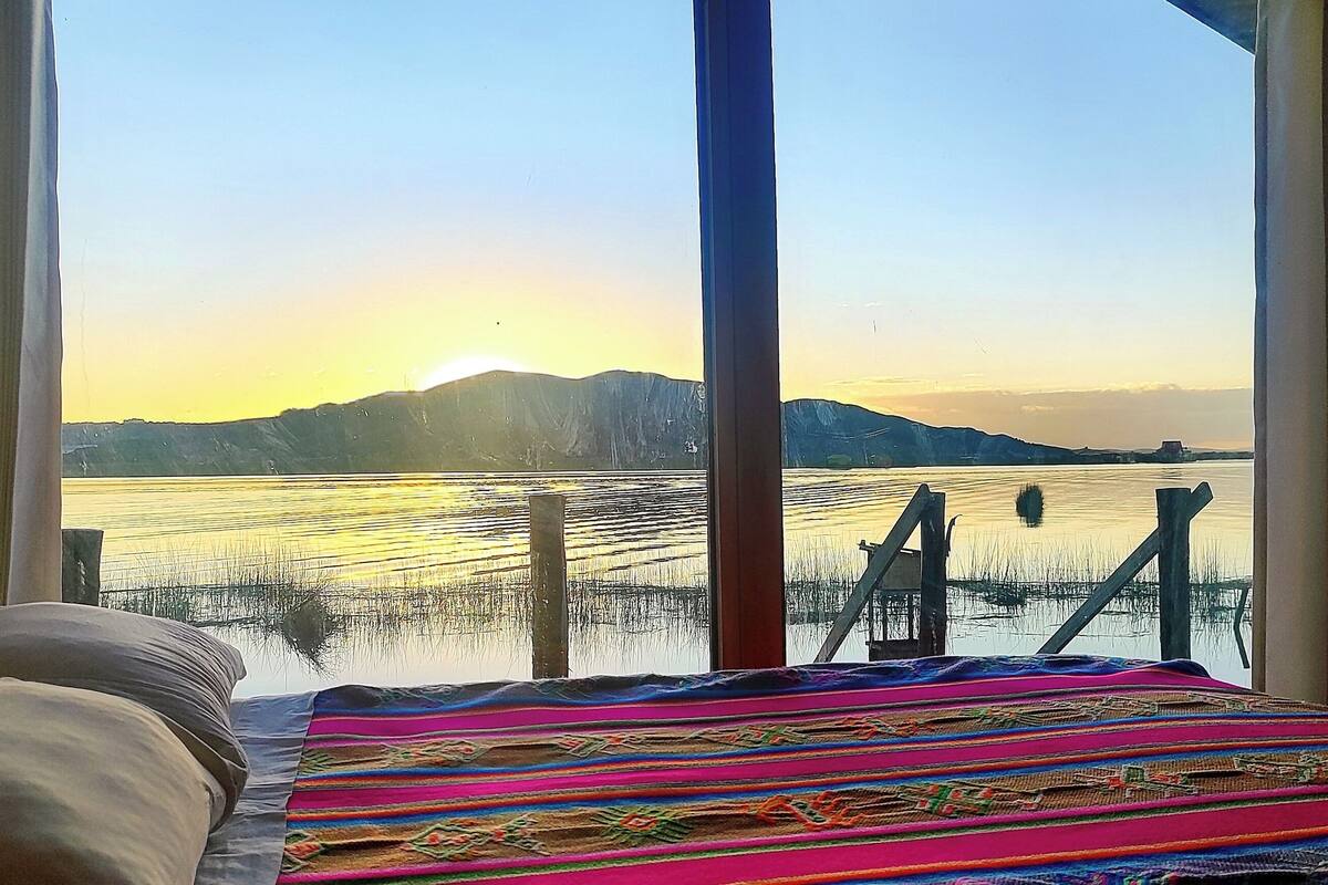 A bed with a colorful blanket, a window with a view of a calm lake and distant mountains during sunset.