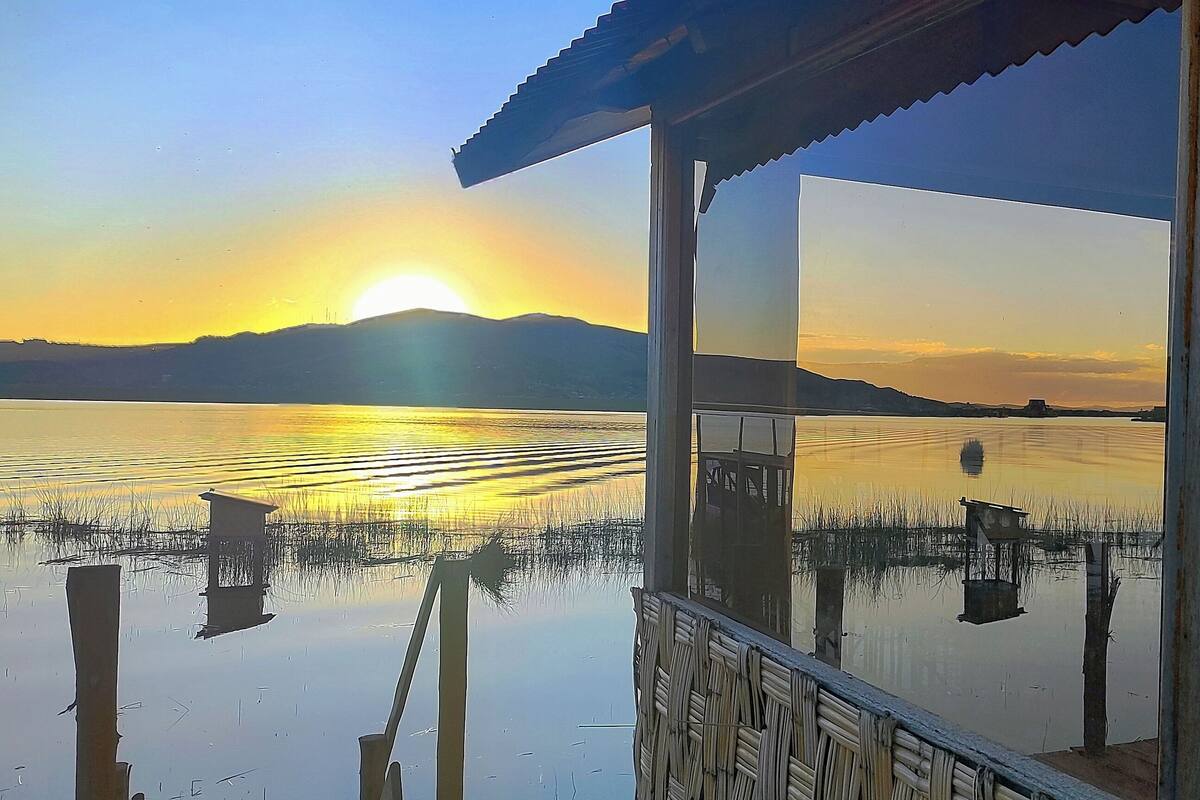 A view from a covered outdoor area with a reflection of the sunset on a calm body of water, surrounded by reeds and a distant mountain range.