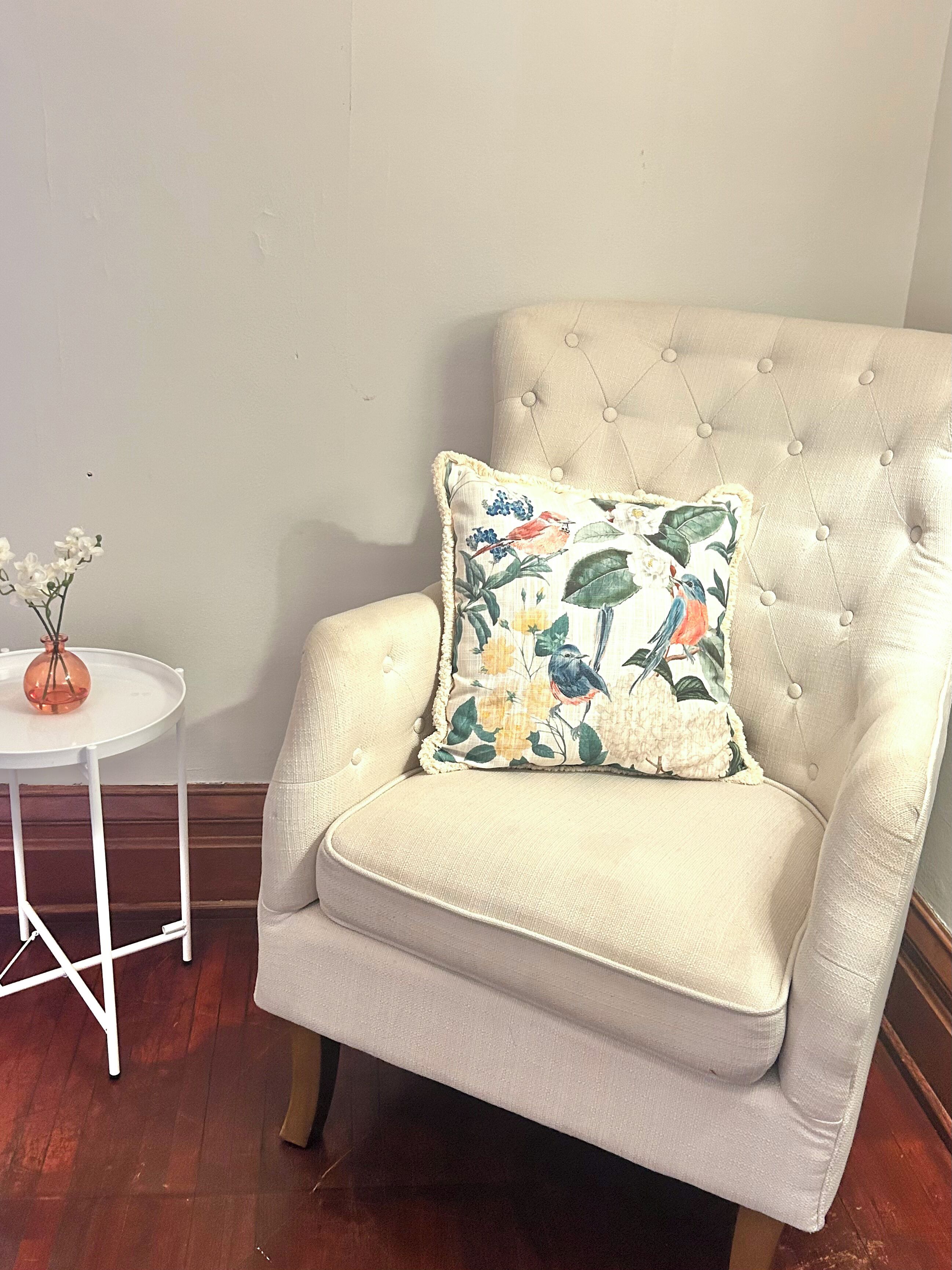 Un sillón tapizado con cojines estampados, una mesita blanca con un florero y suelo de madera.
