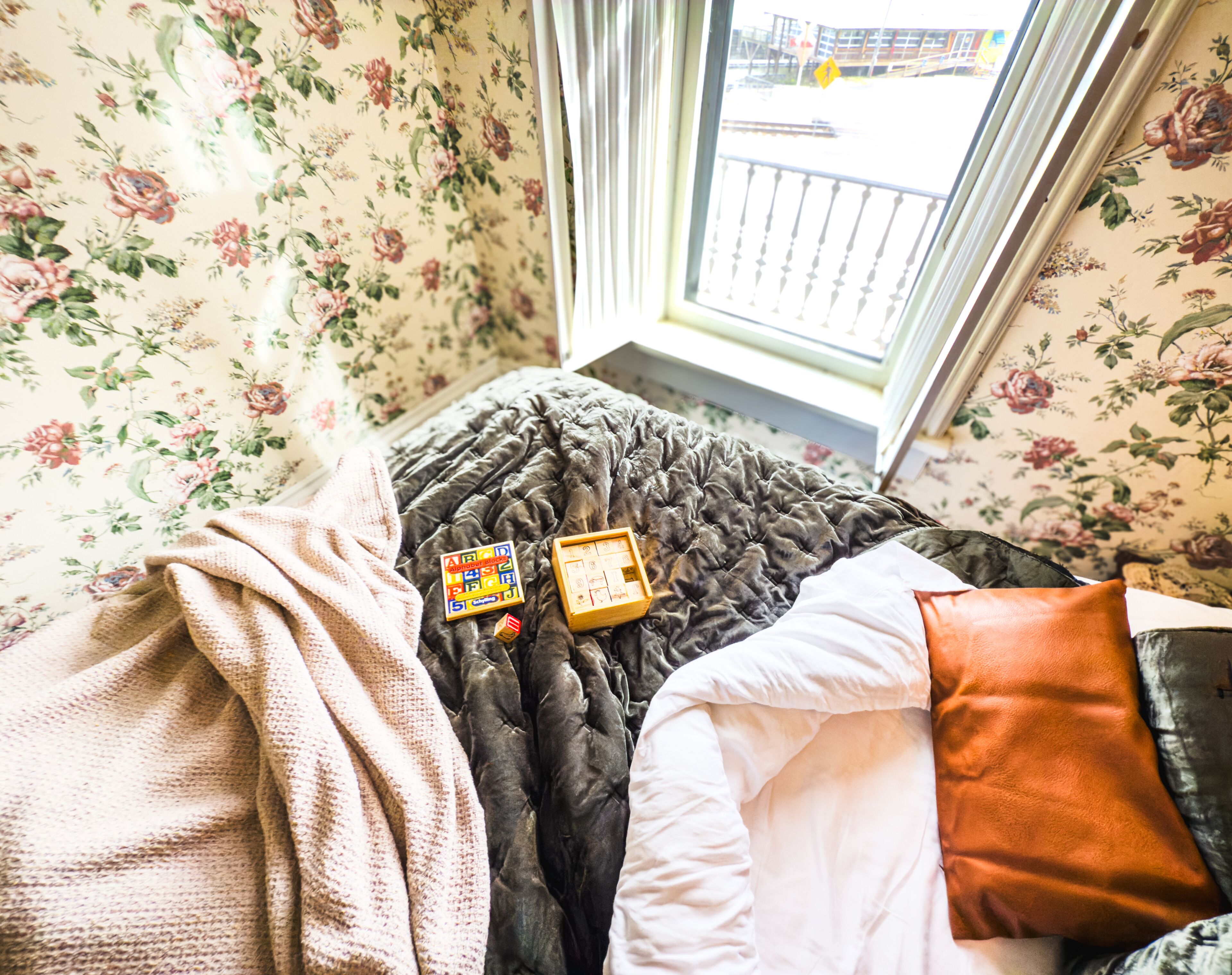 A bed with a floral wallpaper, a window with a view of a building, and two children's toys on the bed.