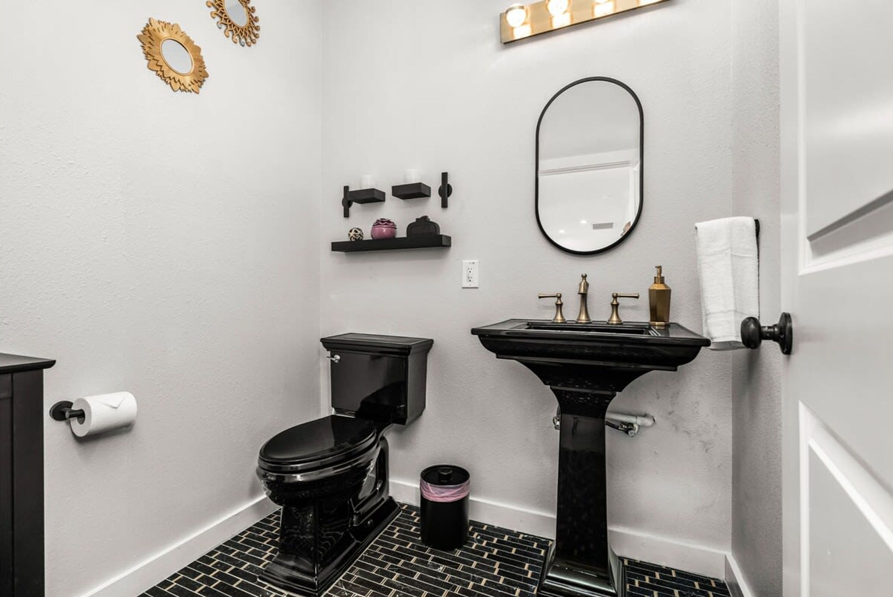 Une salle de bain comprenant des toilettes, un lavabo et un miroir. On y trouve des étagères avec des articles de toilette et une poubelle.
