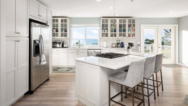 A modern kitchen with white cabinetry, a central island with bar stools, and a view of the ocean through large windows. - Perch - La Fiesta 34: Luxe Retreat w/ Serenity View & Firelit Comfort (Coupeville)