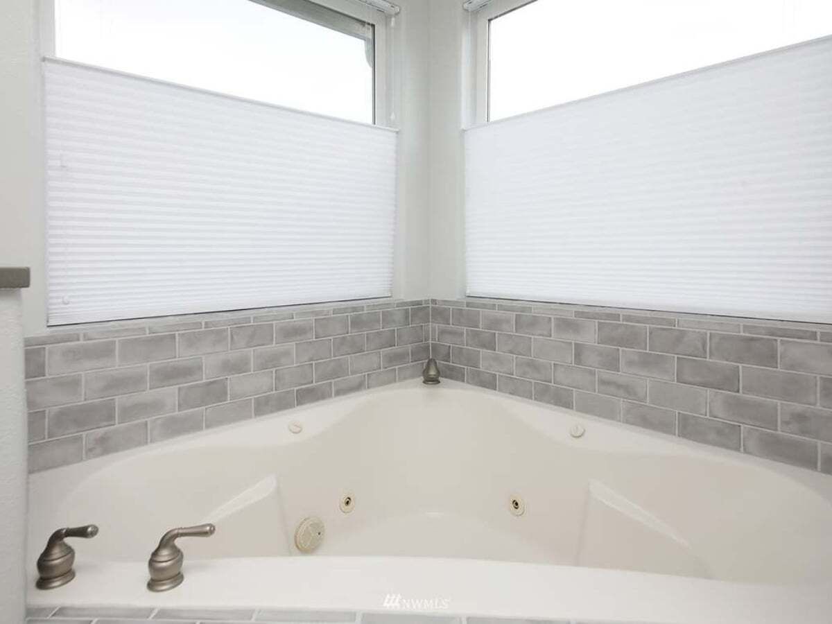 Une salle de bain avec une baignoire blanche, des murs recouverts de carreaux gris et une fenêtre avec des stores.