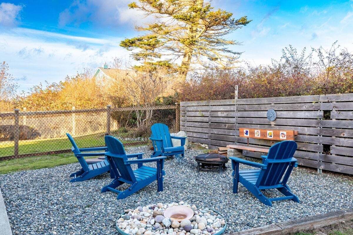 A backyard with a fire pit, three Adirondack chairs, and a wooden fence.