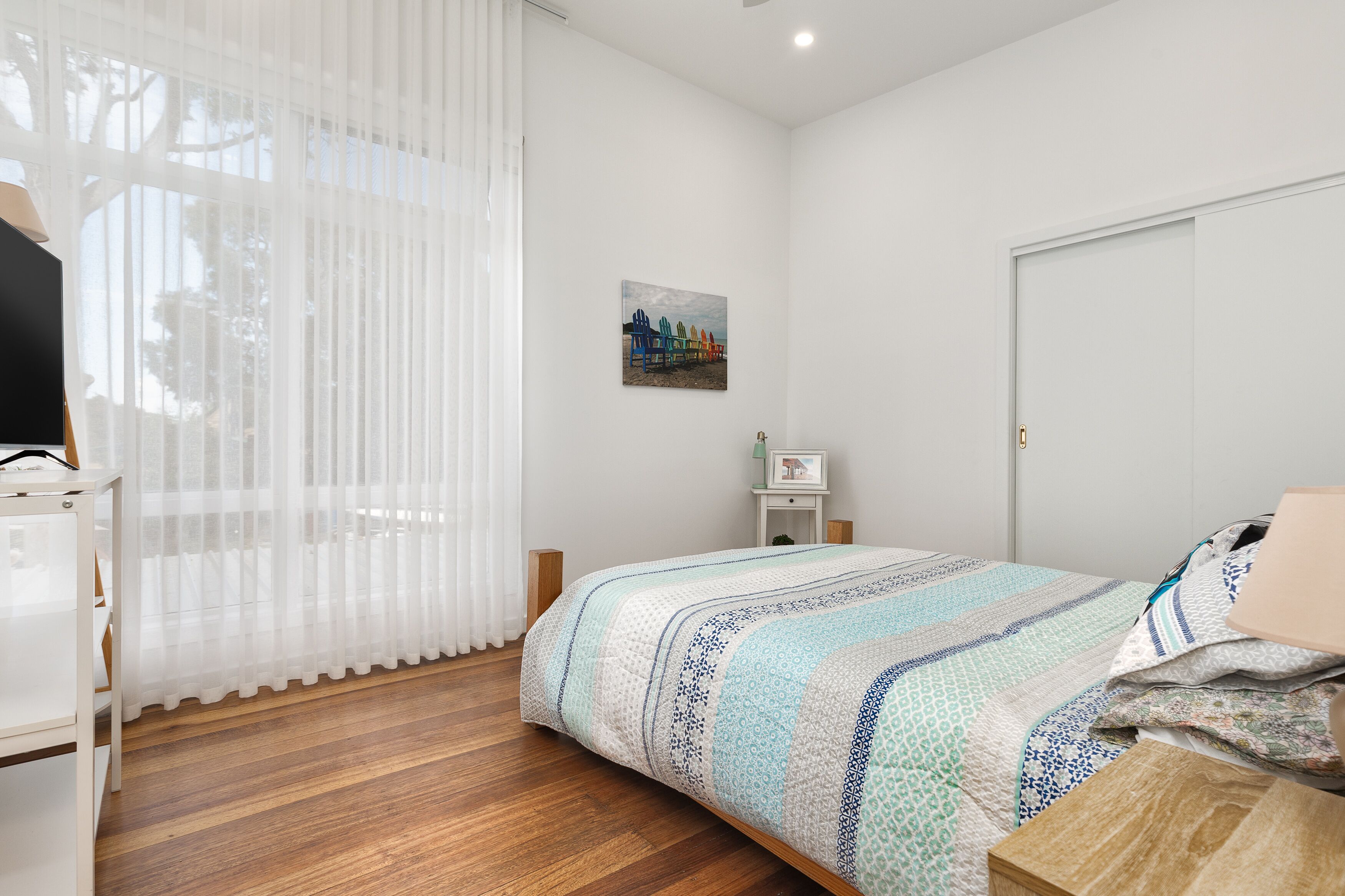 A bedroom with a bed, a bedside table, a nightstand, a window with sheer curtains, and a wall-mounted picture.