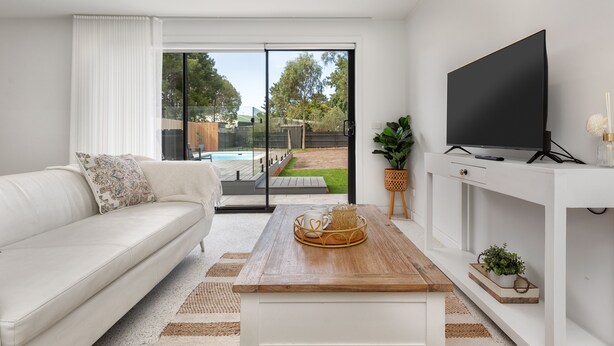 Een moderne woonkamer met een witte bank, een houten salontafel met decoratieve objecten, een flatscreen-tv op een witte standaard en een schuifdeur die leidt naar een buitenruimte.