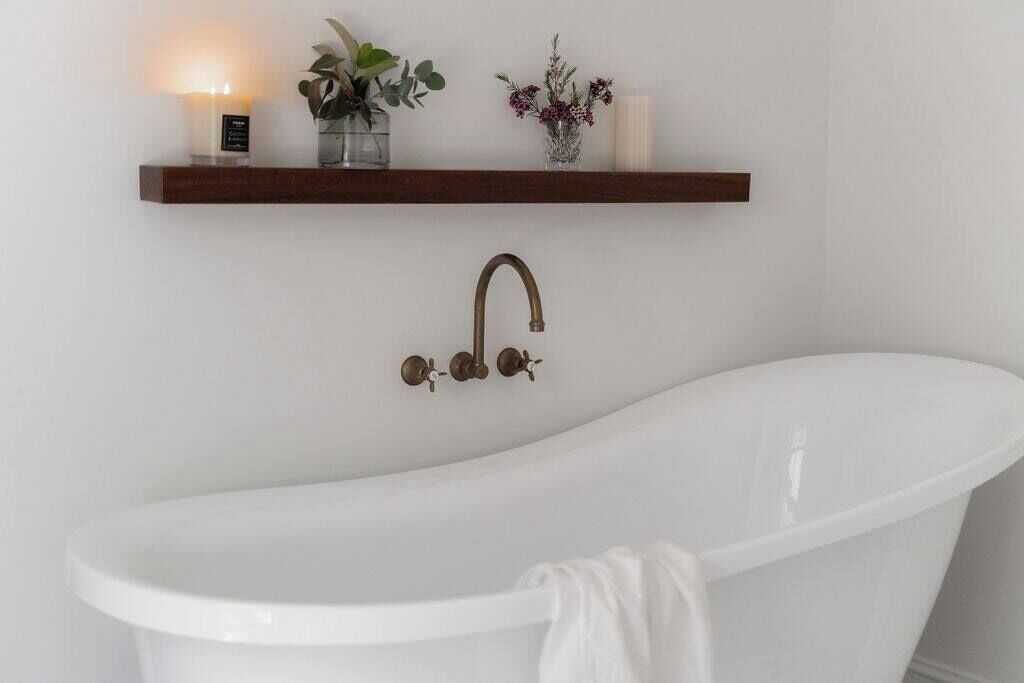 A white bathtub with a brass faucet, a wooden shelf above it holding a candle, a small plant, and a vase with flowers.