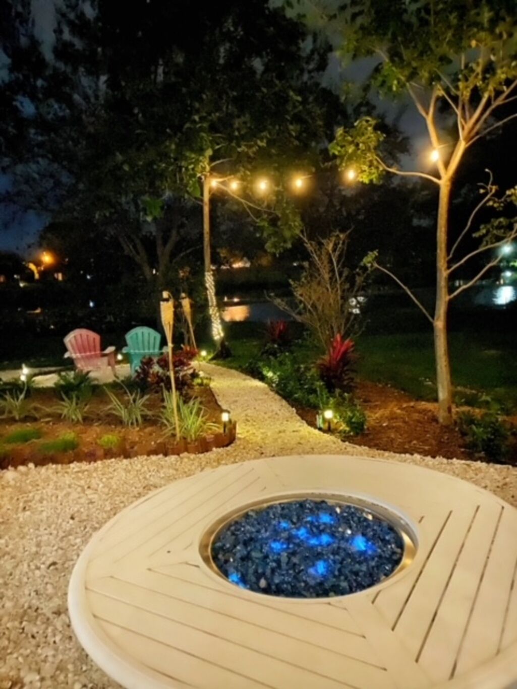 Un jacuzzi rond doté d’un bord en bois et d’une fontaine centrale, entouré d’un chemin de gravier et de parterres de fleurs avec différentes espèces végétales.