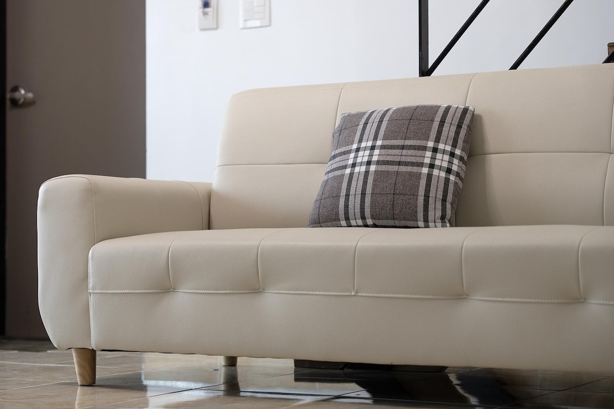 A beige sofa with a plaid pillow, positioned in a room with a tiled floor and a door in the background.