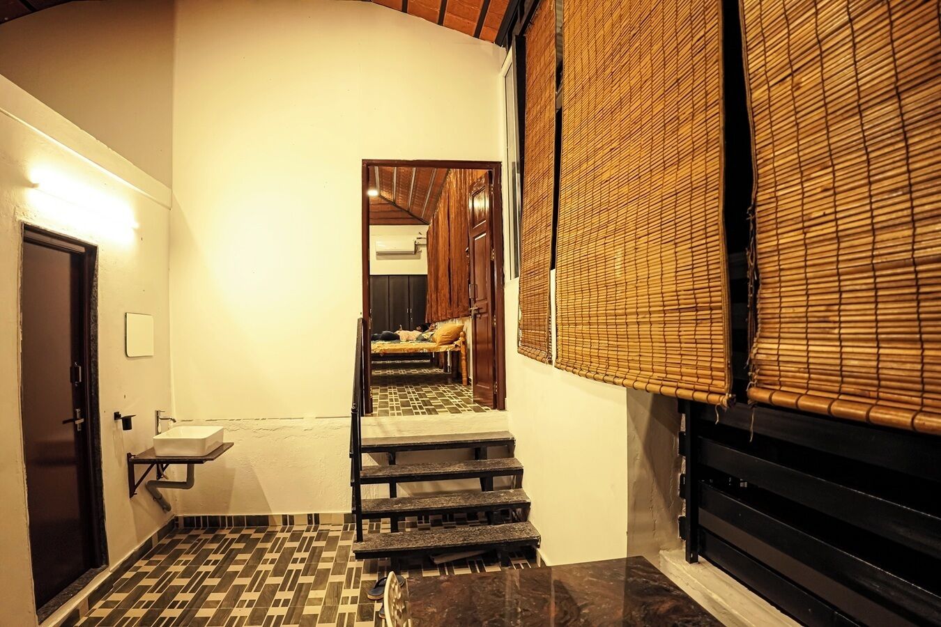 Un pasillo estrecho con suelo de baldosas, un lavamanos pequeño y una escalera de madera que lleva a un dormitorio con cama y mesita de noche.
