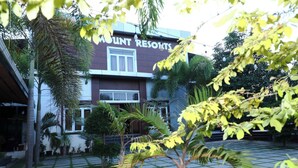 Um edifício com a placa 'Mount Resorts', um pátio pavimentado, palmeiras e outras diversas plantas.