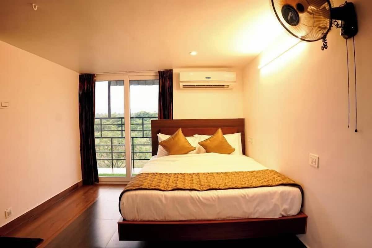 A hotel room with a bed, a ceiling fan, an air conditioning unit, and a view of greenery through the window.