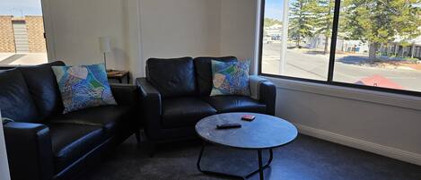 A room with two black leather sofas, a small round table, and a large window with a view of a street and trees.