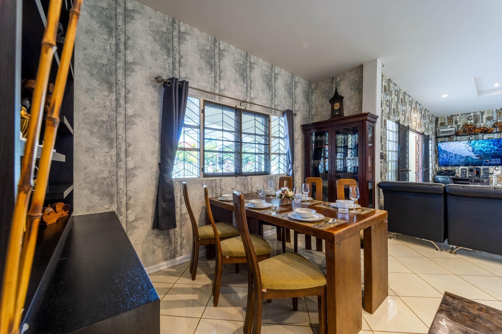 Un comedor con una mesa de madera dispuesta para cuatro personas, rodeada de sillas con asientos acolchados. Se ve una ventana grande con vistas al exterior, y un televisor está montado en la pared.
