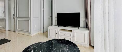 Ein modernes Wohnzimmer mit einem Flachbildfernseher auf einer weißen Kommode, einem Couchtisch aus schwarzem Marmor und einem großen Fenster mit Vorhängen.
