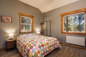 A bedroom with a large bed, two windows, a nightstand with a lamp, a radiator, and a framed picture on the wall.