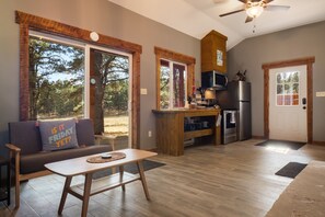 Cocina moderna con refrigerador, microondas y estufa, zona de comedor con mesa y sillas, y ventanas amplias con vistas a árboles.