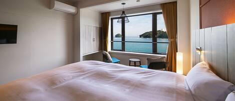 A hotel room with a large bed, a window with a view of the sea, a small table, and a chair.