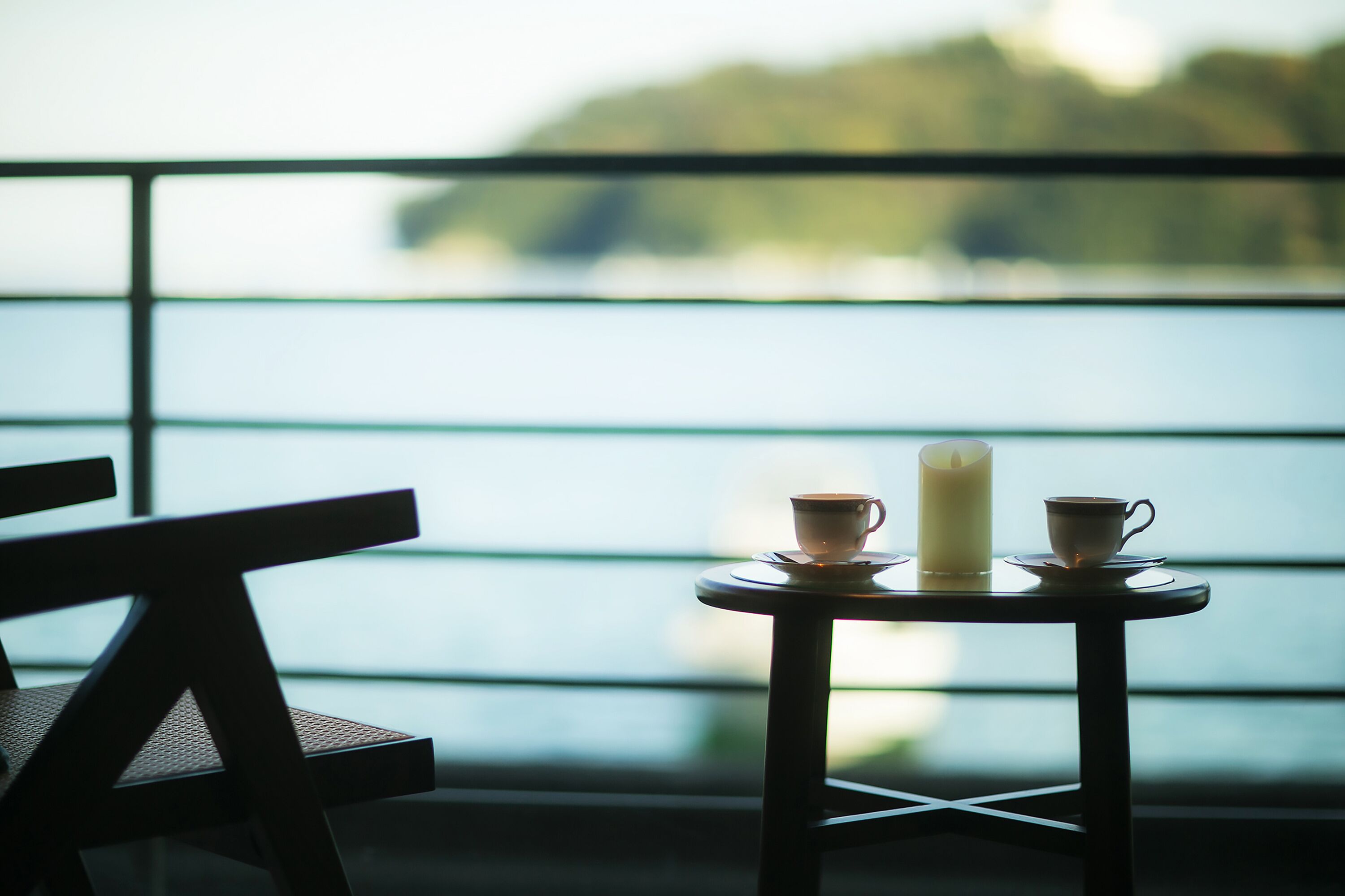 A table with two cups and a candle, set against a window with a view of a body of water.