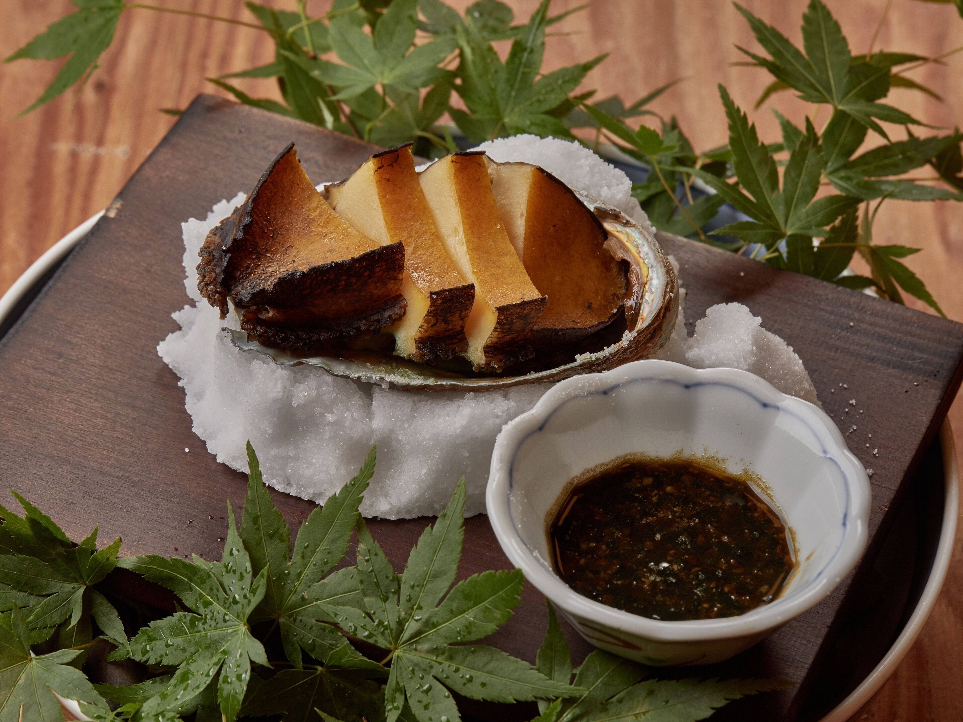 Un plat composé d’un morceau de viande posé sur un lit de glace, accompagné d’un petit bol de sauce, le tout présenté sur un plateau en bois décoré de feuilles vertes.