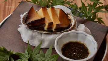 Un plat composé d’un morceau de viande posé sur un lit de glace, accompagné d’un petit bol de sauce, le tout présenté sur un plateau en bois décoré de feuilles vertes.