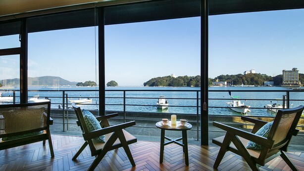 Un balcón con suelo de madera, dos sillones de mimbre, una mesita redonda con una vela, y vistas al mar con barcos y edificios a lo lejos.