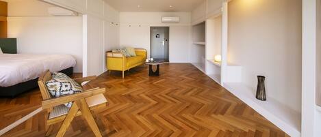 A modern hotel room with a wooden herringbone floor, a bed, a yellow sofa, a small table, and a chair.