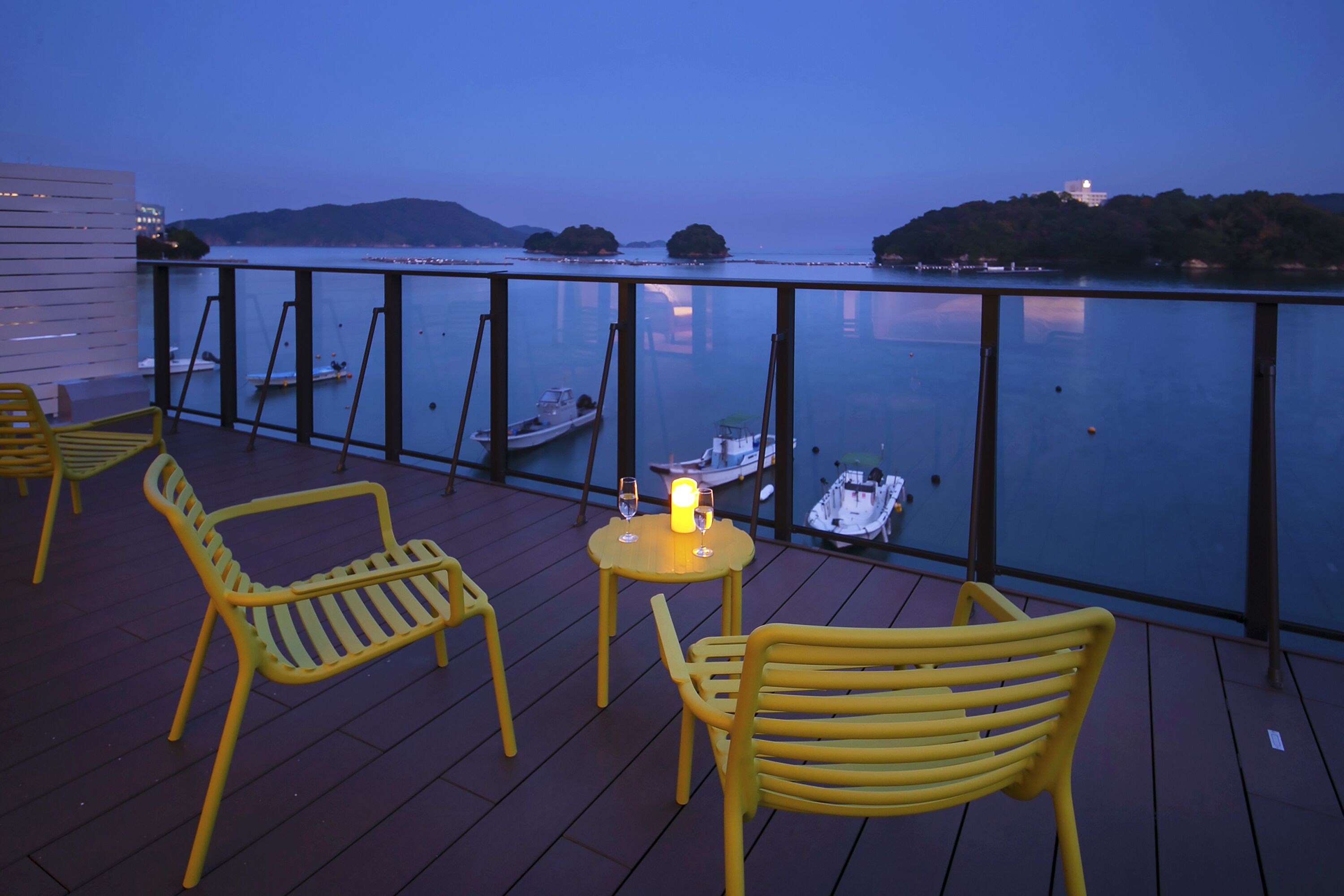 A deck with yellow chairs and a table, overlooking calm water with boats and distant hills.