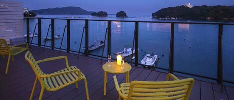 A deck with yellow chairs and a table, overlooking calm water with boats and distant hills.