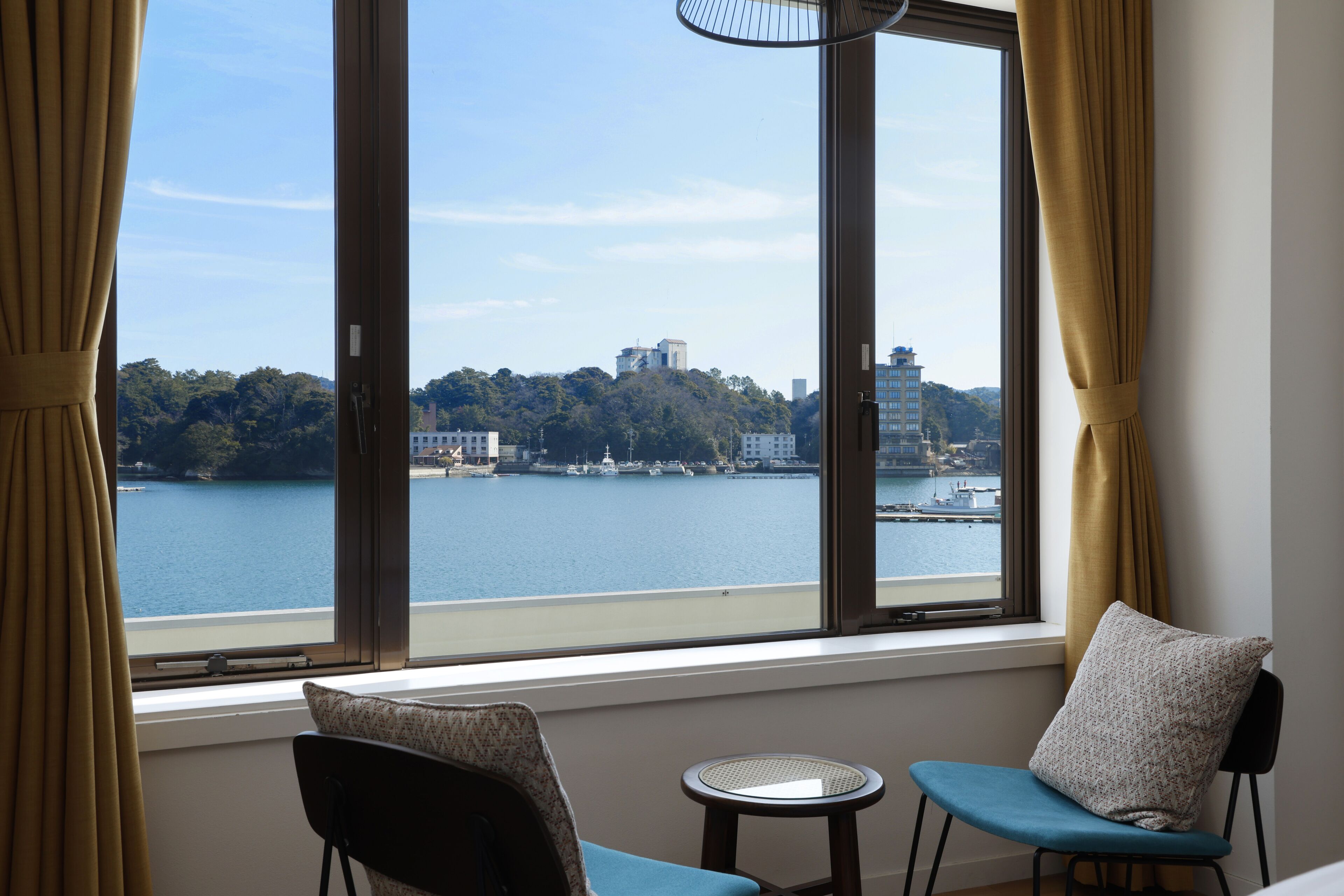 A room with a large window overlooking a body of water and a hilly landscape. There is a chair and a small round table in the room.