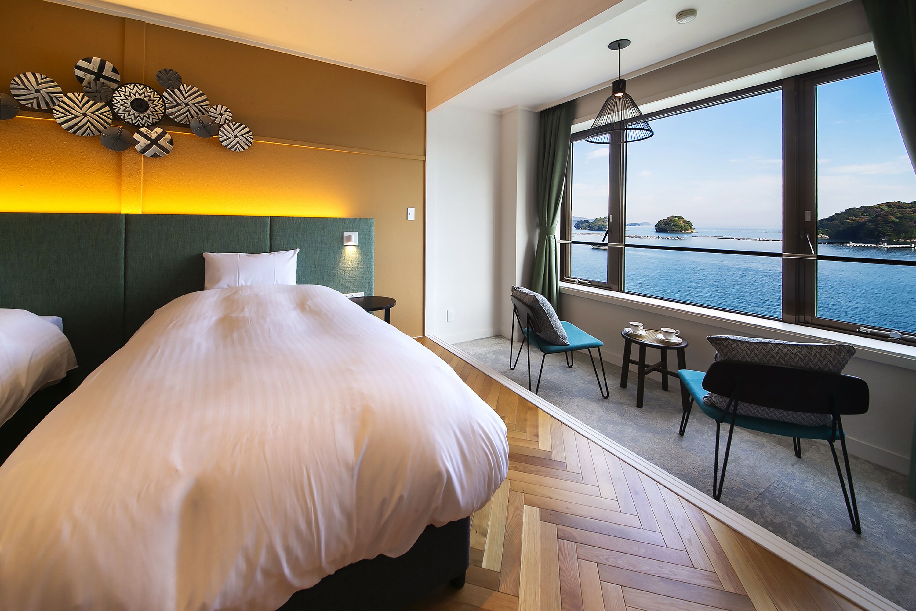 Une chambre d’hôtel avec un grand lit, une vue sur la mer, un coin salon avec une table et des chaises, et un élément décoratif mural.