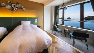 A hotel room with a large bed, a view of the sea, a seating area with a table and chairs, and a decorative wall piece.