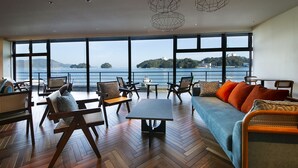 Amplia habitación con un gran ventanal con vistas a una masa de agua y una isla distante. Varias áreas de descanso, suelo de madera y mobiliario moderno.
