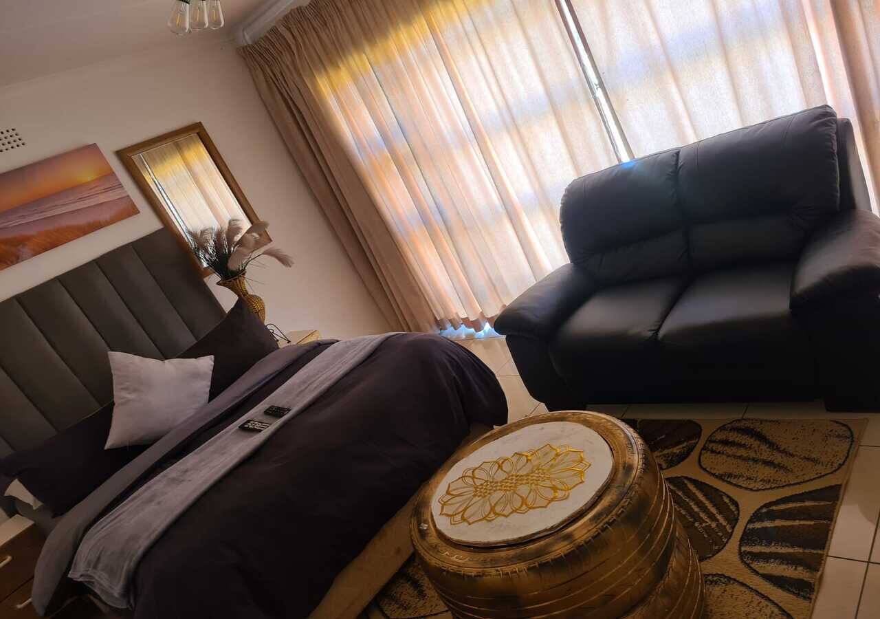 A bedroom with a bed, a headboard, a bedside table, a lamp, a mirror, a window with curtains, and a sofa.