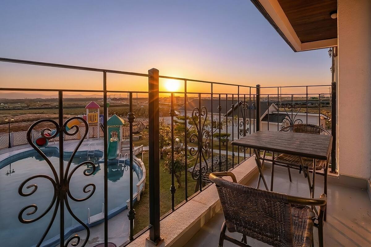 A balcony with a view of a swimming pool and playground, a table and chair, and a sunset in the background.