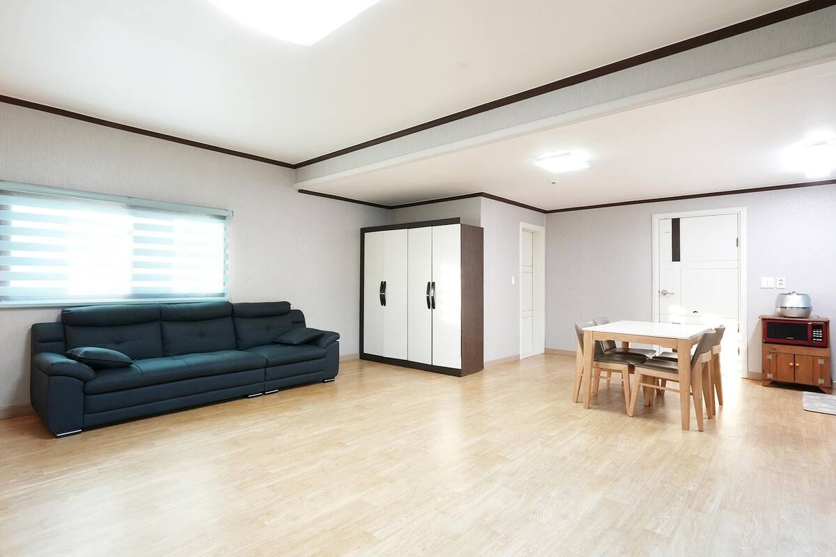 A modern living room with a sofa, a dining table with chairs, a wardrobe, and a window with blinds.