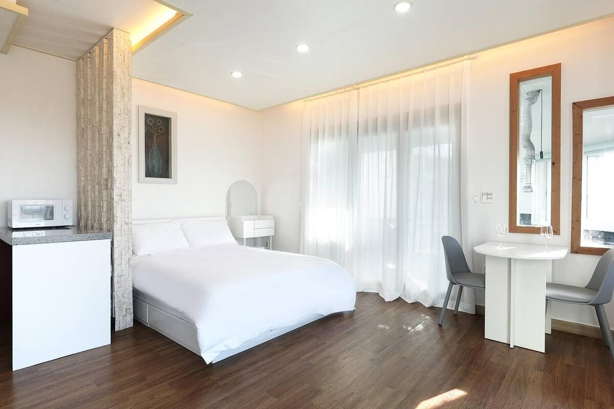 A modern hotel room with a bed, a desk, a chair, a microwave, and a large window with curtains.