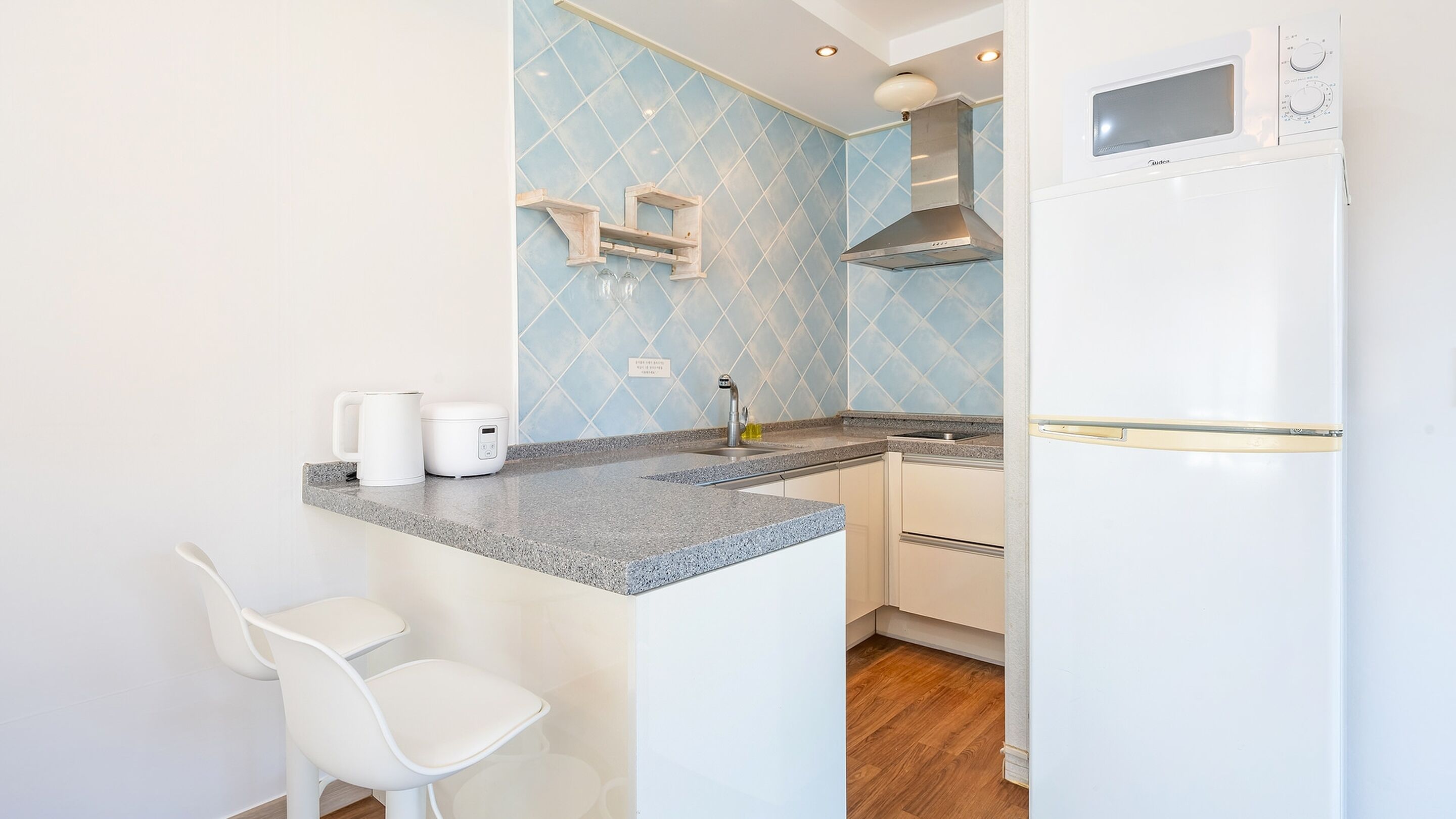 A compact kitchen with a granite countertop, a built-in microwave, and a stainless steel range hood. There are two white bar stools and a small island with a kettle and a rice cooker.
