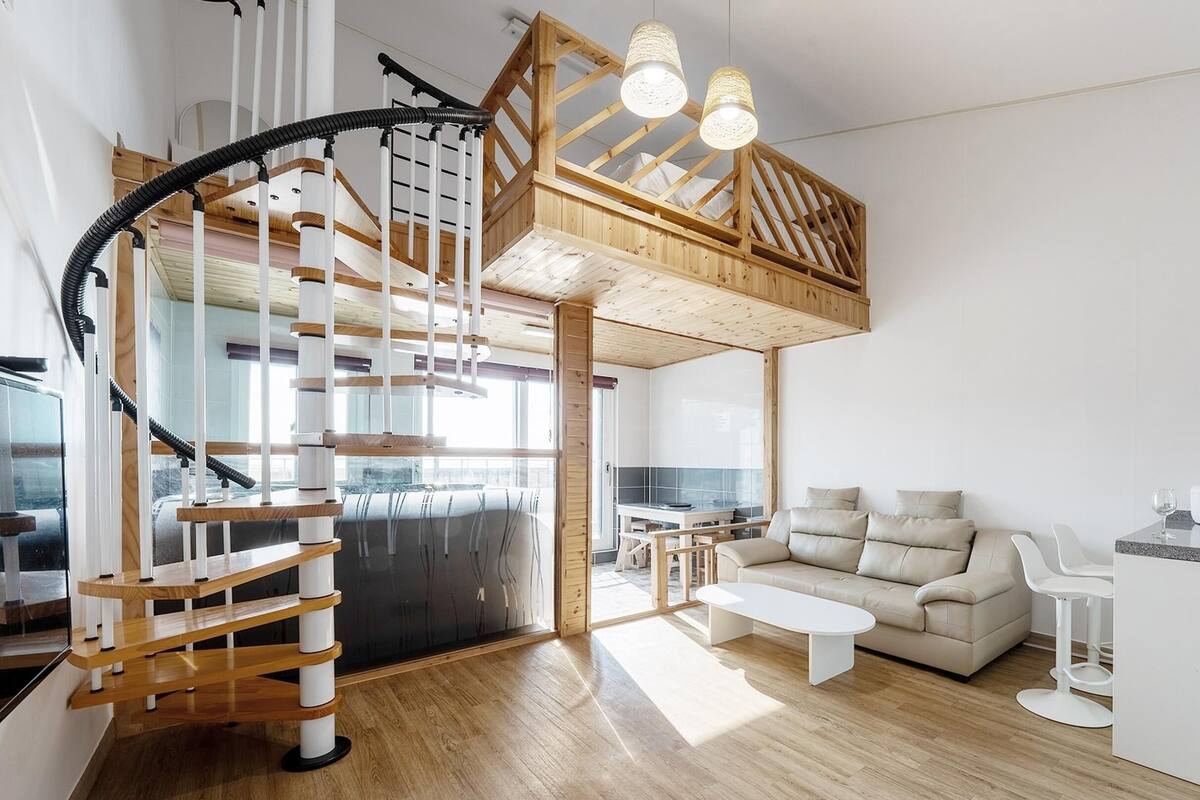 A modern interior with a wooden staircase, a loft bedroom, a living area with a sofa, and a kitchen with a coffee machine.