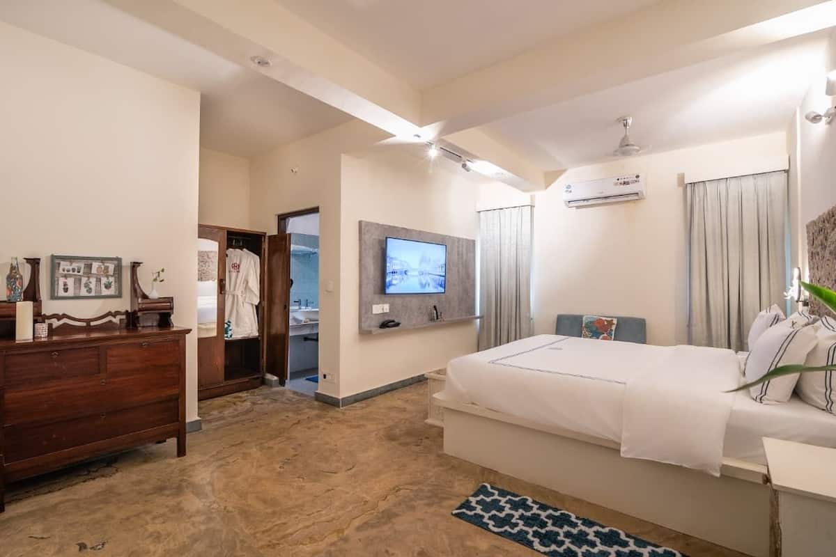 A modern hotel room with a large bed, a flat-screen TV mounted on the wall, a wooden dresser, and a bathroom visible through an open door.