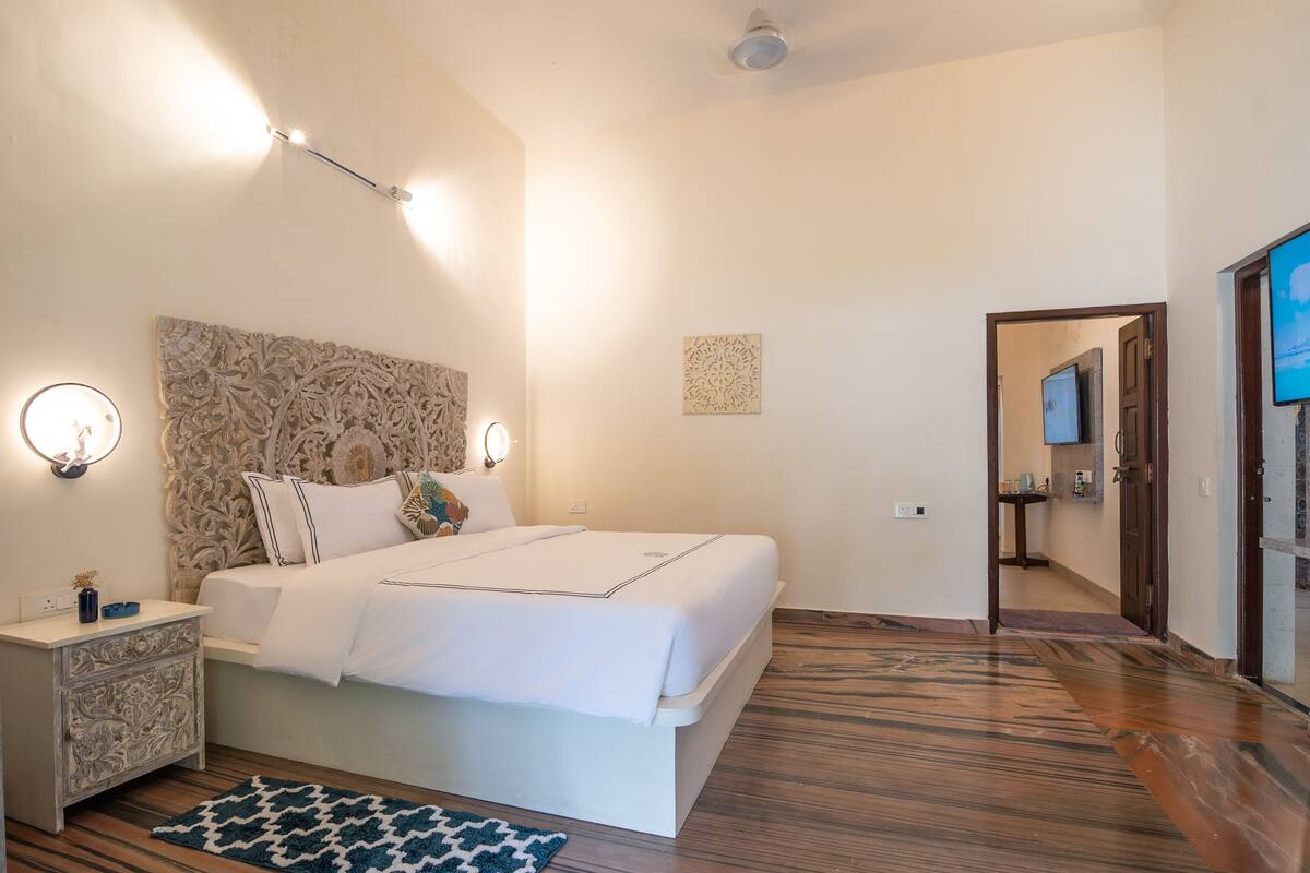A bedroom with a large bed, bedside lamps, a decorative headboard, and a wooden floor.