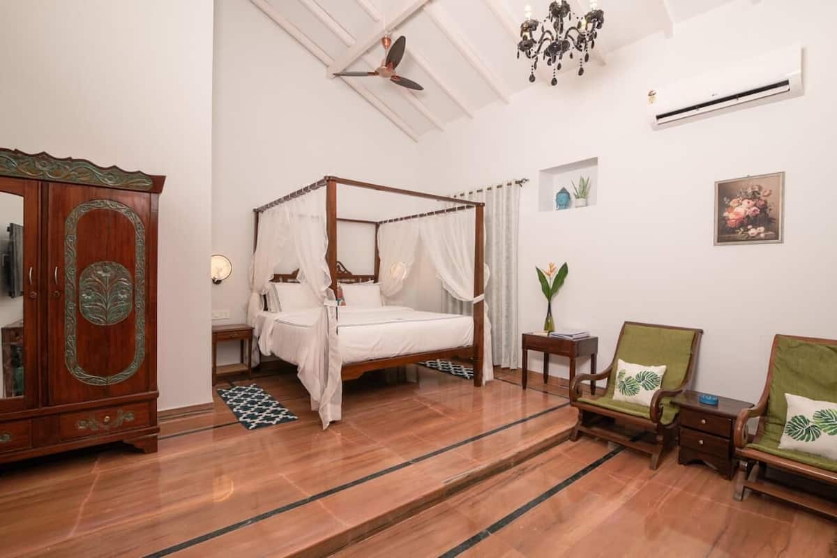 A bedroom with a four-poster bed, a wooden wardrobe, two chairs, a small table, and a ceiling fan.