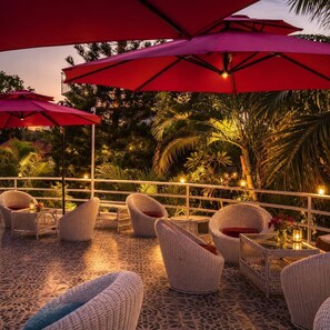 Un espacio al aire libre con sillas y mesas de mimbre, sombrillas rojas y vistas a follaje tropical.