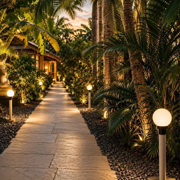 A pathway lined with palm trees and illuminated by spherical lights, leading to a building with a thatched roof.