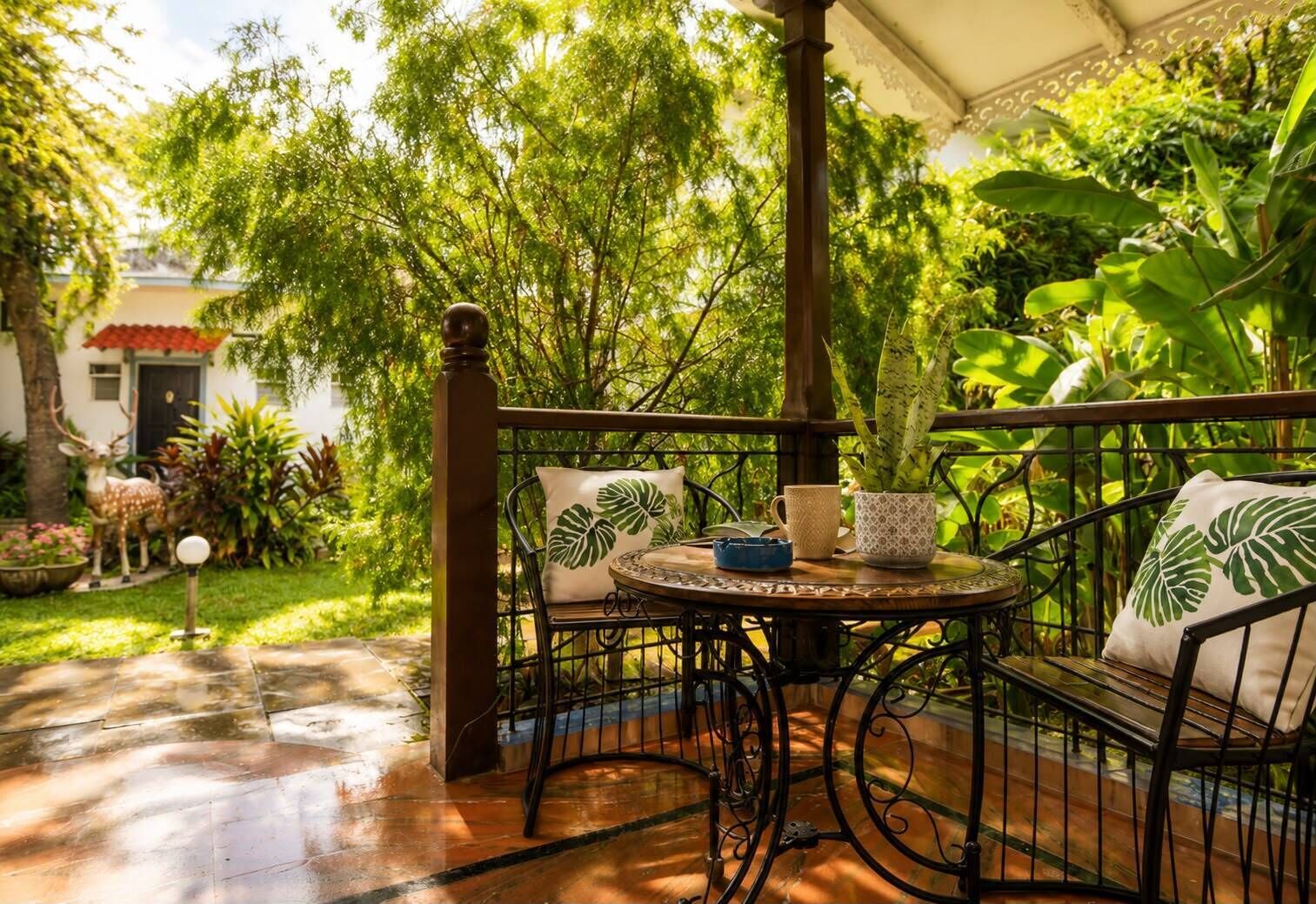 A covered outdoor seating area with a table and chairs, surrounded by lush greenery and a garden.