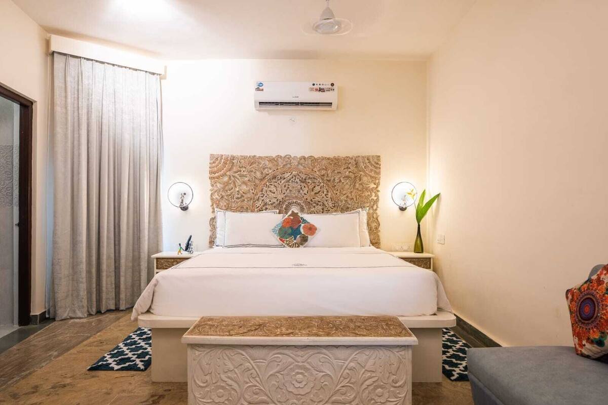 A hotel room with a large bed, an air conditioning unit, a decorative headboard, a bedside table with a lamp, and a small bench.