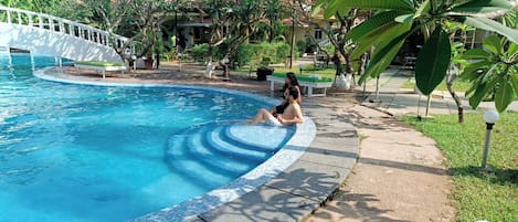 A swimming pool with a white bridge, surrounded by greenery and a paved walkway.